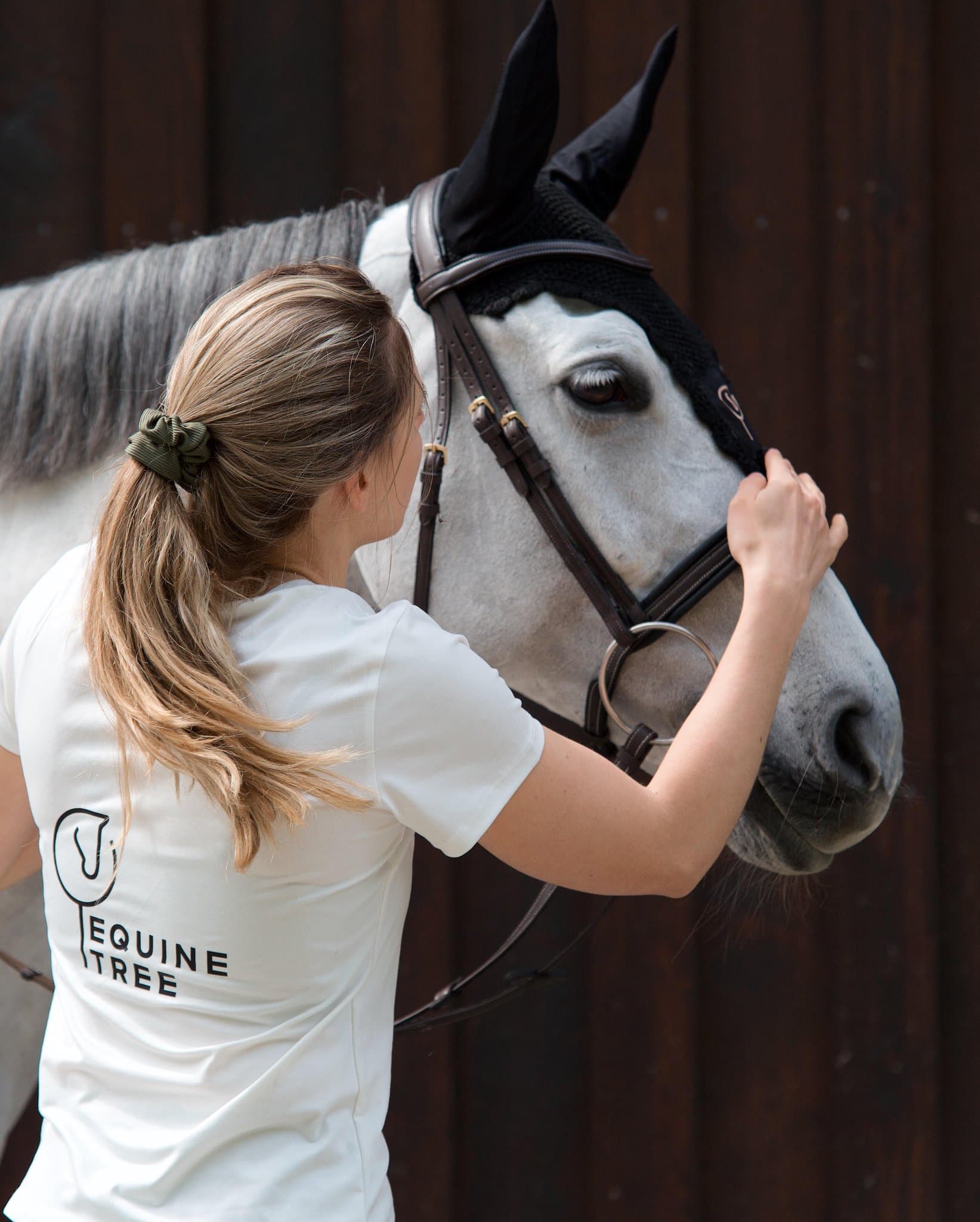 Rider in an Equinetree shirt adjusting the bridle on a grey horse